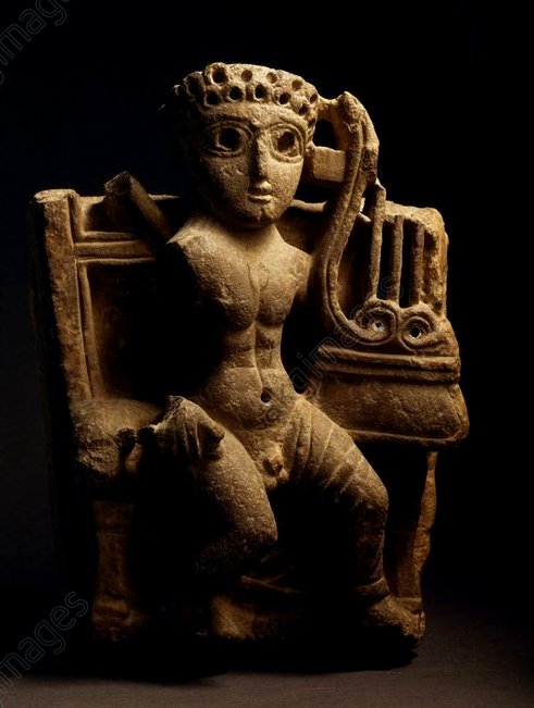 A damaged limestone sculture of a seated naked man with large eyes, curled hair, and a lyre in his left hand. 