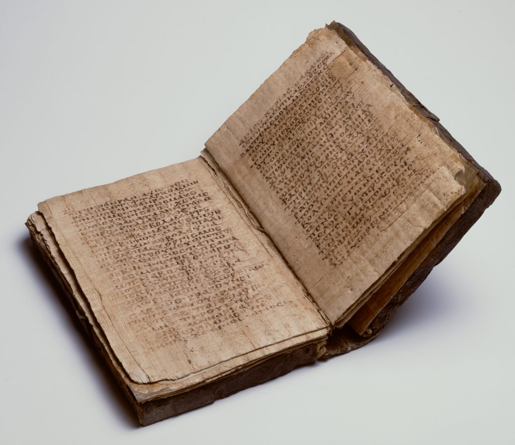 An image of an ancient codex (book) with a slightly damaged dark brown leather cover. Here the book is open to show the papyrus pages, with dense Coptic text on each side.