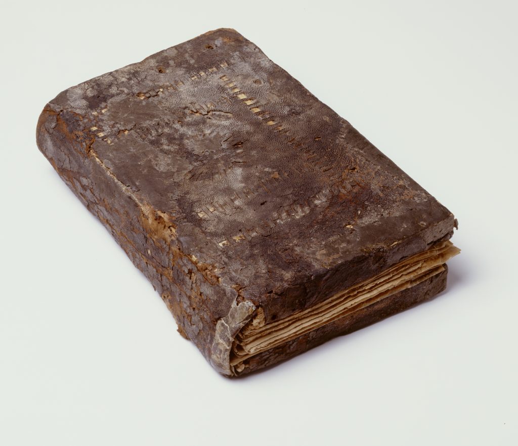 An image of an ancient codex (book) with a slightly damaged dark brown leather cover. Some decoration in the form of concentric rectangles with dotted lines is visible on the front. The papyrus pages are just visible although the book is closed.
