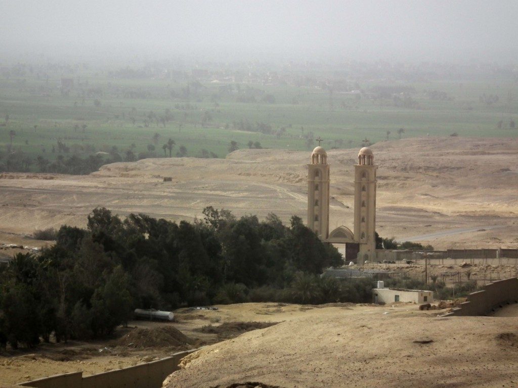 A picture of a monastery with two towers in the Egyptian desert; in the foreground are a group of trees beside the monastery, while in the background the cultivated land of the Fayum can be seen.
