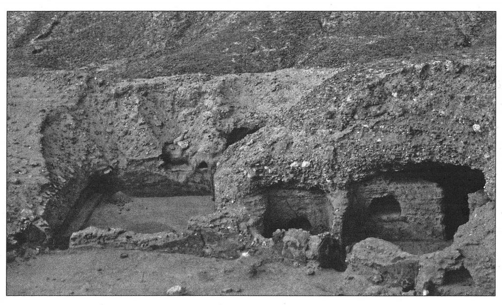 Black and white photograph of the remains of a monastery cut into the rock of a mountain, with walls and roofs supplemented by mud-brick.