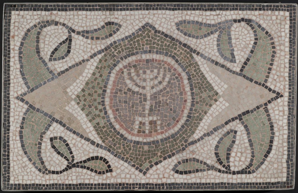 A mosaic showing a six-branched menorah with three legs within a diamond shape, decorated by curling plant-like forms.