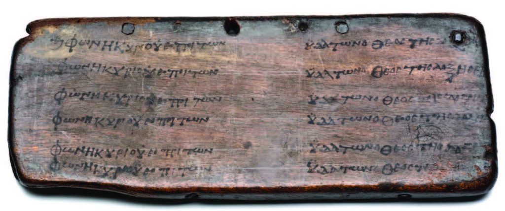 A dark wooden tablet, with Greek writing in two columns and four holes drilled into its upper part.