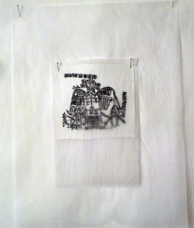 An image of a printing of a magical drawing of an angel surrounded by magical signs; the printed image, on white paper, is pinned to a wall through three larger pieces of tracing paper, and covered by one piece of tracing paper, through which is it still clearly visible.