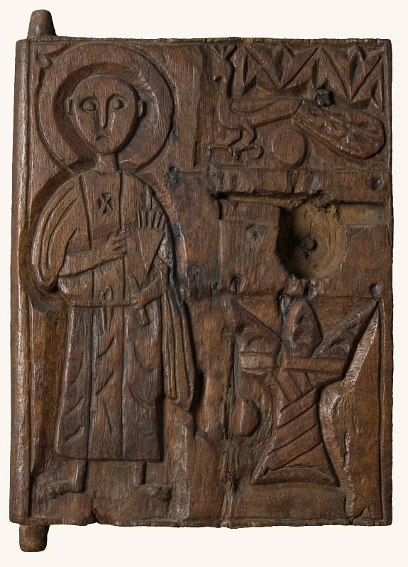 A wooden panel with tenons at the top and bottom left showing it was used as a door or lid. On the left is carved a figure in a monastic robe with a halo. On the upper right is a peacock, and on the lower right is a horned altar.