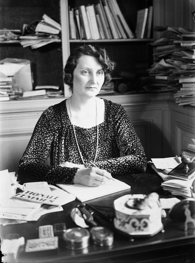 Black and white photo of a woman sitting at a desk and writing with a pen. In the background can be seen shelves piled with books and papers.