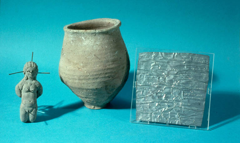 An image on a blue background. On the left is a clay figure of a naked woman with curly hair tied back and a thick necklace; she is on her knees and with her hands tied behind her back, and nails can be seen piercing her eyes, ears, head and genitals. To the figure's right is a small clay jar, and further right is a lead sheet held in a glass case. It has writing on it, and has been folded many times.