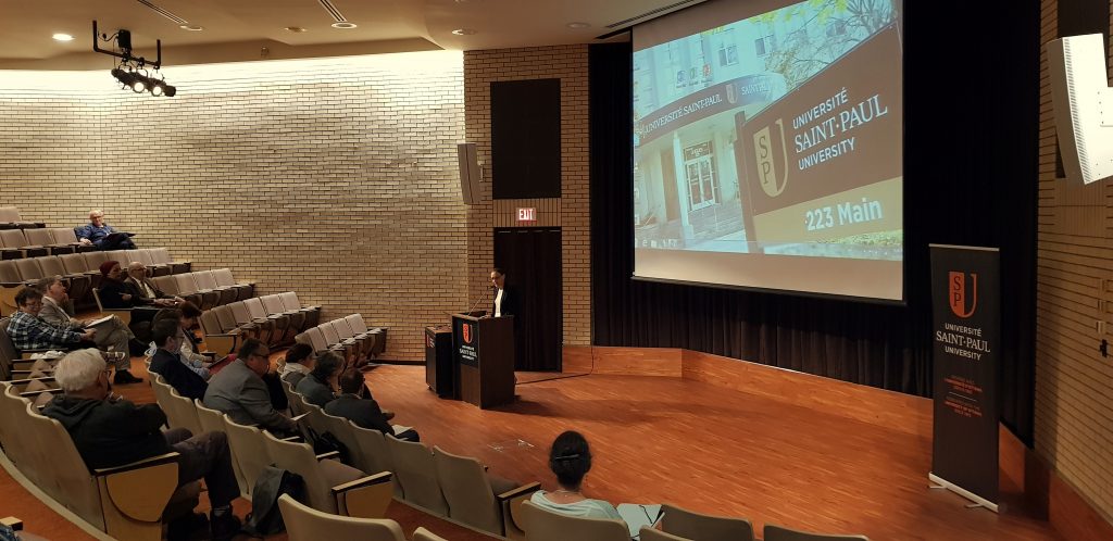 A photo of an amphitheatre, with a dozen people visible sitting on the seats. A woman in a black suit is speaking at the podium, and on the screen in the background a photo of the entrance to the University of Saint Paul, with a sign in front of it clearly visible giving its name.